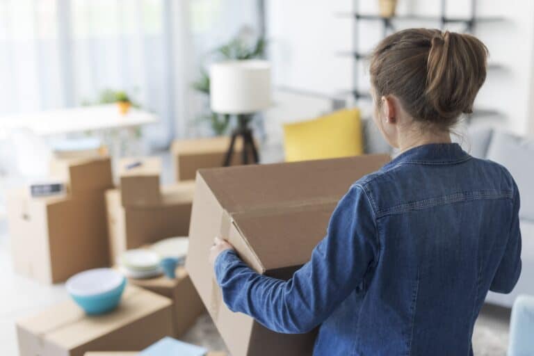 A woman is carrying a sealed box to her living room full of packed boxes in the concept of moving to a new house in Orpington.