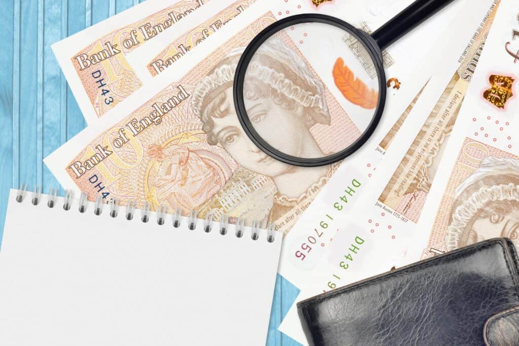 A magnifying glass on top of a British pound bill with a notebook and wallet in the concept of 'What Are the Hidden Costs in Home Removals Services'.